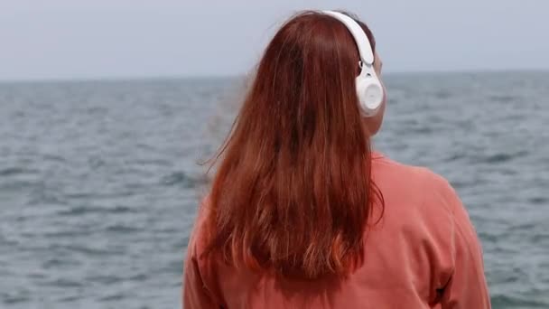 Vista posterior de la mujer que usa auriculares escuchando música o podcast desde la aplicación de teléfonos inteligentes contra el mar — Vídeo de stock