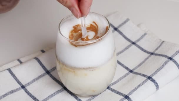 Donna mano mescolando caffè latte con caramello con cucchiaio di vetro sul tavolo bianco — Video Stock