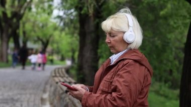 Portrait of adult senora 50 years old woman using smartphone and listening music by headphones on street outdoors