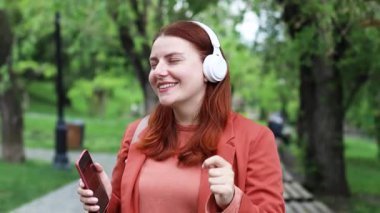 Smiling young caucasian millennial girl holding smartphone using mobile application, dancing, singing, fooling around, having fun, technology outdoors in city park.