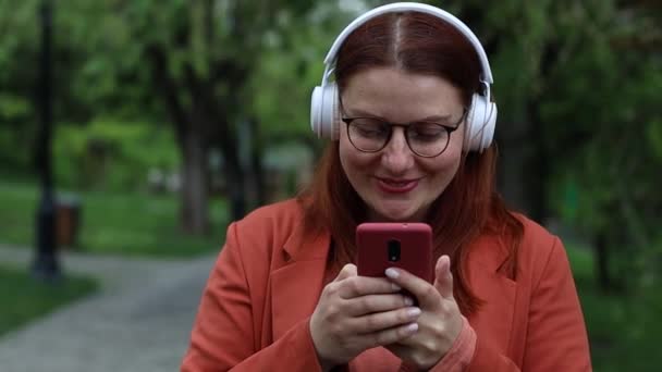 Young Caucasian millennial woman wearing glasses holds smartphone using mobile apps outdoors. Happy young person in white wireless headphones listening to music — стоковое видео