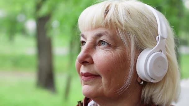 Caucasian happy 50 years old woman wearing white headphones listening to music with eyes closed on street outdoors. — Stockvideo