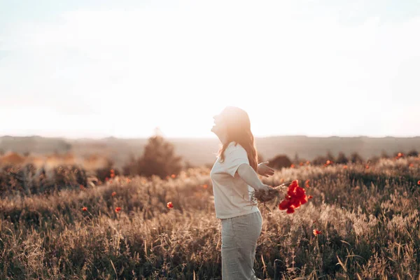 Incrível jovem linda menina ruiva com buquê de flores em um campo de papoula ao pôr-do-sol. Conceito de liberdade — Fotografia de Stock