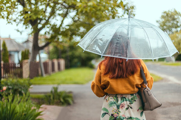 Back view of young woman in bright clothes under a transparent umbrella in bad weather in the city park
