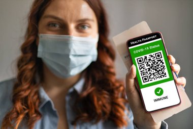 Pasaport, bilet kartı ve dijital sağlık uygulamalı akıllı telefon taşıyan maske takan bir gezgin kadın. Covid-19 Sağlık Pasaportu.