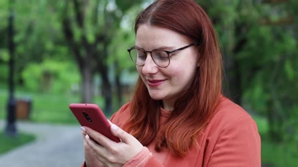 Smil kaukasisk kvinne i briller med smarttelefon ved hjelp av mobilteknologi utendørs. Gledelig ung kvinne som tekster for å sjekke sosiale nettverk i byparken. – stockvideo