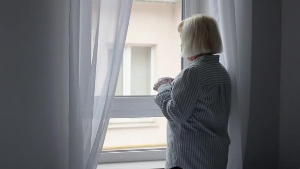 Een volwassen blonde vrouw staat bij het raam en drinkt warme koffie of thee drinken, geniet van een vroege rustige ochtend — Stockvideo