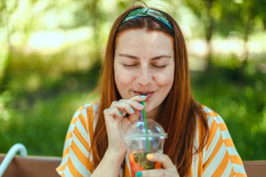 Şehir parkındaki ahşap bankta oturmuş soğuk kokteyl içen neşeli yaz kadını dinleniyor. Sağlıklı gıda konsepti