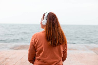 Kulaklık takan, müzik dinleyen ya da denize karşı akıllı telefon uygulamasından podcast yapan seksi saçlı kadının arka planı.