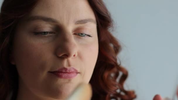 Gros plan portrait de femme caucasienne aux cheveux bouclés positif applique un maquillage à l'aide d'une brosse à poudre sur le mur gris — Video