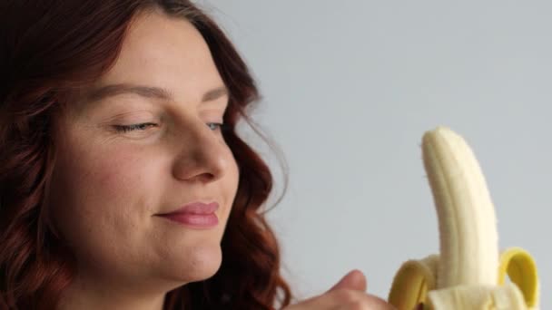 Close up de alegre mulher caucasiana encantadora mordendo banana madura amarela e olhando para a câmera. Cara feminina comer banana — Vídeo de Stock