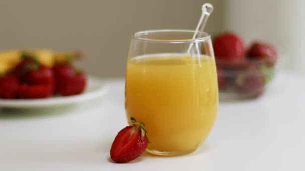 Склянка свіжовичавленого апельсинового соку з полуницею на білому кухонному столі — стокове відео