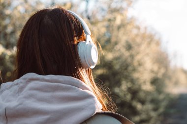 Güneşli bir günde şehir parkında yürürken kulaklıkla müzik dinleyen beyaz bir genç kadın.