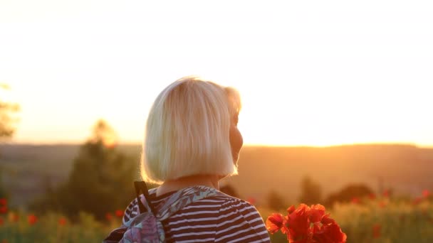 Adulto donna caucasica turista con zaino e papavero mazzo di fiori al tramonto nel campo. vacanze estive. Ragazza felice con i capelli biondi godendo il sole. — Video Stock