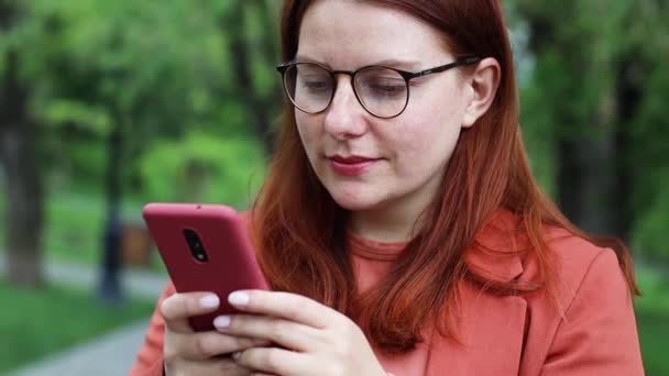İş kadını akıllı telefonu dışarıda mobil uygulama teknolojisi kullanarak tutuyor. Şehir parkındaki sosyal medya uygulamalarına bakıp mesajlaşan mutlu genç bir kadın.. — Stok video