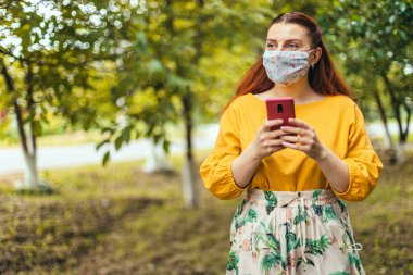 Coronavirus koruyucu tıbbi maskeli mutlu kız şehir parkında cep telefonu kullanıyor.