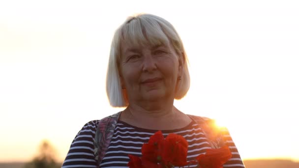 Eine erwachsene kaukasische Touristin mit Rucksack und Mohnblumenstrauß bei Sonnenuntergang auf dem Feld. Sommerferien. Glückliches Mädchen mit blonden Haaren genießt die Sonne. — Stockvideo