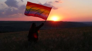Gün batımında çiçek açan bir tarlada parlak LGBT bayrağı taşıyan özgür genç bir kız. Rüzgarda el sallayan gökkuşağı bayrağı