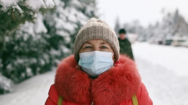 Eine erwachsene Seniorin in medizinischer Schutzmaske blickt im Winterpark in die Kamera. Soziale Distanzierung während der Coronavirus Covid 19 Pandemie — Stockvideo