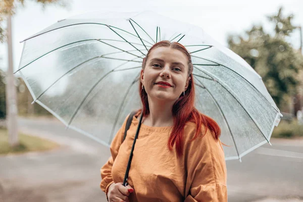 Ευτυχισμένη νεαρή γυναίκα με φωτεινά ρούχα κάτω από μια διάφανη ομπρέλα σε κακές καιρικές συνθήκες στο πάρκο της πόλης — Φωτογραφία Αρχείου