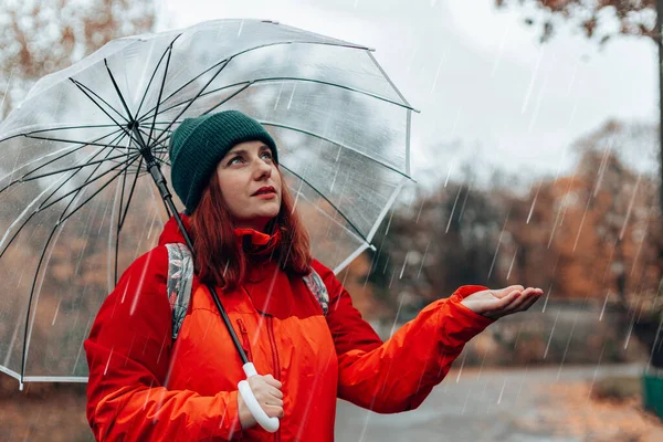 Fehér nő hátizsákkal és piros kabátban sétál nedves esős utcákon egy átlátszó esernyő alatt — Stock Fotó