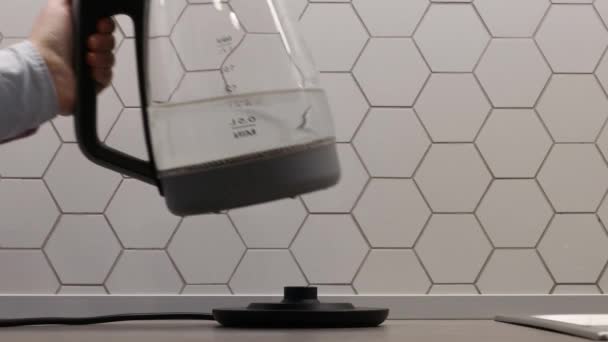 Κοντινό πλάνο ενός γυναικείου χεριού βάζει ένα διαφανές ηλεκτρικό βραστήρα στο περίπτερο πατώντας το κουμπί διακόπτη ρεύματος. Διάφανο ηλεκτρικό βραστήρα με βραστό νερό στο τραπέζι στην κουζίνα — Αρχείο Βίντεο
