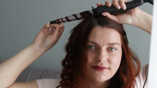 Piękna dziewczyna z długimi czerwonymi włosami zrobić jej fryzurę z suszarką żelaza — Wideo stockowe