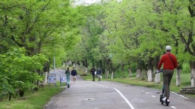 Two young teen girls ride an electric scooter on the asphalt road in city park. Back view