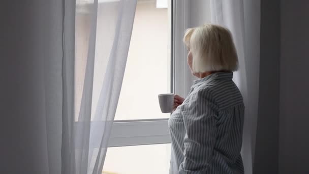 Morning of adult blonde woman stands by the window and drinks hot coffee or tea drink, enjoys an early quiet morning — Wideo stockowe