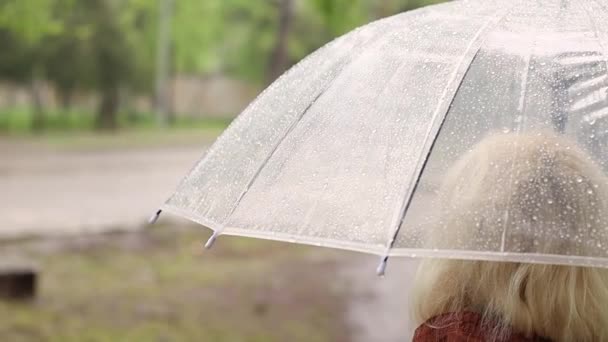 Vista trasera de una mujer rubia bajo paraguas transparente durante la lluvia, mal tiempo, aguacero fangoso — Vídeo de stock