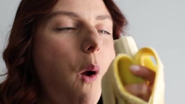 Sarı, olgun muzu ısıran ve kameraya bakan neşeli, beyaz bir kadın. Kadın yüzü muz yiyor.