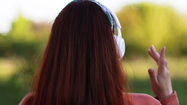 Menutup pandangan dari belakang kepala wanita yang bahagia di usia seribu tahun di headphone di taman kota pada musim panas — Stok Video