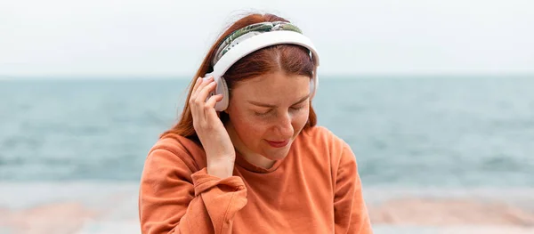 Belle jeune femme caucasienne écoutant de la musique avec des écouteurs le jour ensoleillé tout en marchant près de la mer — Photo