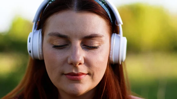 Belle femme caucasienne avec les yeux fermés écoutant de la musique à l'aide d'écouteurs sans fil et smartphone dans le parc de la ville — Photo