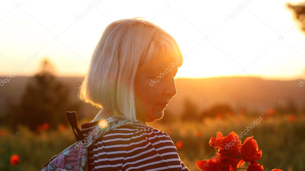 Adult Caucasian woman tourist with backpack and poppy flower bouquet at sunset in the field. Summer vacation. Happy girl with blond hair enjoying the sun.