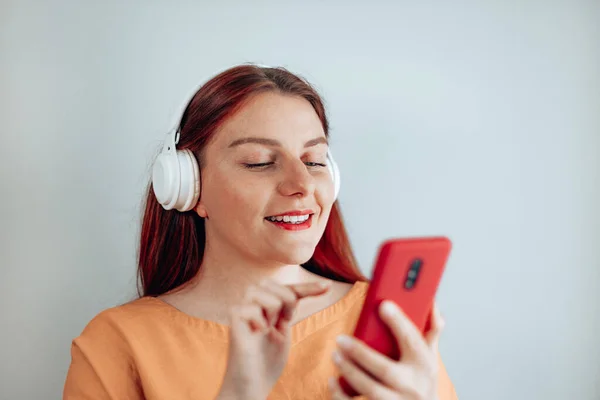 Shot usmívající se mladá žena na sobě bezdrátové bílé sluchátka pomocí svého mobilního telefonu těší stylové moderní hudební melodie — Stock fotografie