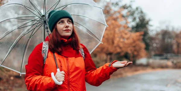 Θετική Καυκάσια γυναίκα με σακίδιο και κόκκινο μπουφάν περπατά σε βρεγμένους βροχερούς δρόμους κάτω από διάφανη ομπρέλα. Φθινοπωρινή βόλτα — Φωτογραφία Αρχείου
