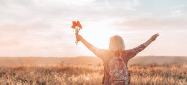 Gün batımında sırt çantalı ve gelincik çiçekli beyaz kadın turistin arka görüntüsü. Turizm, seyahat ve sağlıklı yaşam tarzı konsepti.