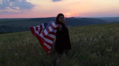Gün batımında arkası dönük mutlu bir kadın. Elleri açık, ABD bayrağı taşıyor.