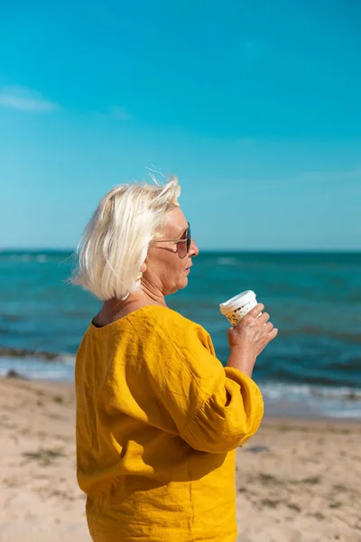 Vuxen blond kvinna i solglasögon och en gul blus dricker kaffe från en pappersmugg på stranden en solig dag. Kaffe att ta med — Stockfoto