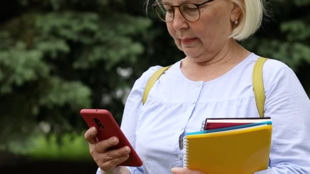 Une enseignante adulte portant des lunettes avec des livres et des cahiers utilise un smartphone alors qu'elle se trouve dans la rue dans un parc de la ville. — Video