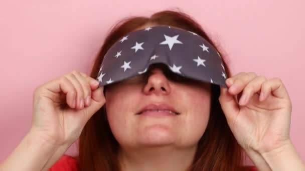 Sleepy young caucasian girl puts on a sleep mask on her eyes on a pink wall background. — Stock Video