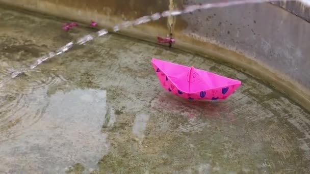 El niño pone un barco de papel rosa brillante en el agua y lo empuja lejos. Niños divirtiéndose junto al agua. — Vídeos de Stock