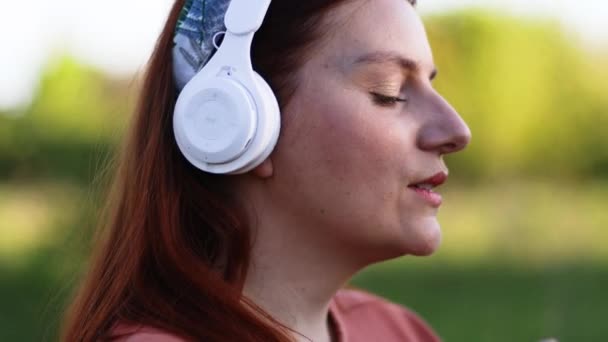 Blank gelukkig vrouw dragen witte hoofdtelefoon luisteren naar muziek met ogen dicht op straat buiten. — Stockvideo