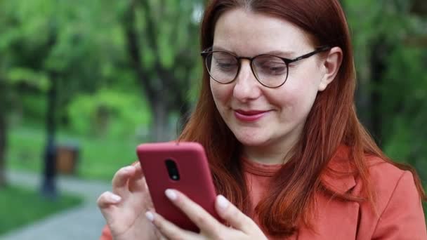 Menina millennial caucasiano jovem em óculos segurando smartphone usando a tecnologia de aplicação móvel ao ar livre. Jovem feliz mensagens de texto verificando aplicativos de redes sociais on-line no parque da cidade. — Vídeo de Stock