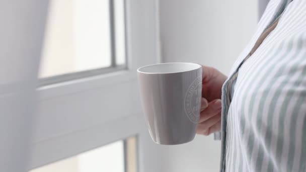 Femme blanche mains tenant une tasse de café chaud à côté de la fenêtre après le réveil le matin à l'intérieur d'une chambre — Video