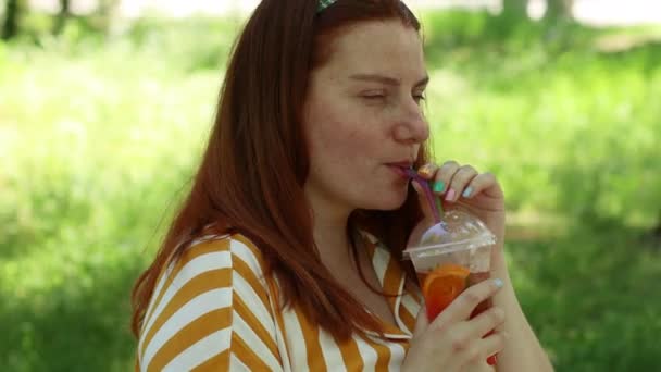 Приваблива довгошерста молода щаслива жінка п'є коктейль, сидячи на лавці в парку, літні канікули — стокове відео