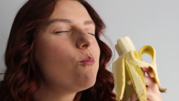 Close up de alegre mulher caucasiana encantadora mordendo banana madura amarela e olhando para a câmera. Cara feminina comer banana — Vídeo de Stock