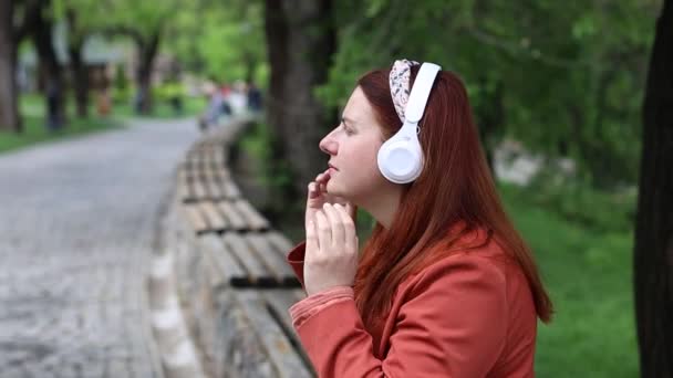 Souriant jeune fille millénaire caucasienne en blanc écouteurs sans fil écouter de la musique en plein air. — Video