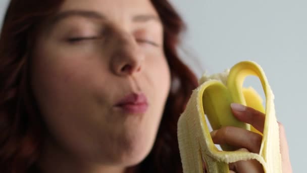 在欢乐迷人的高加索女人的近照下，她咬着黄色成熟的香蕉，看着镜头。吃香蕉的女性脸 — 图库视频影像